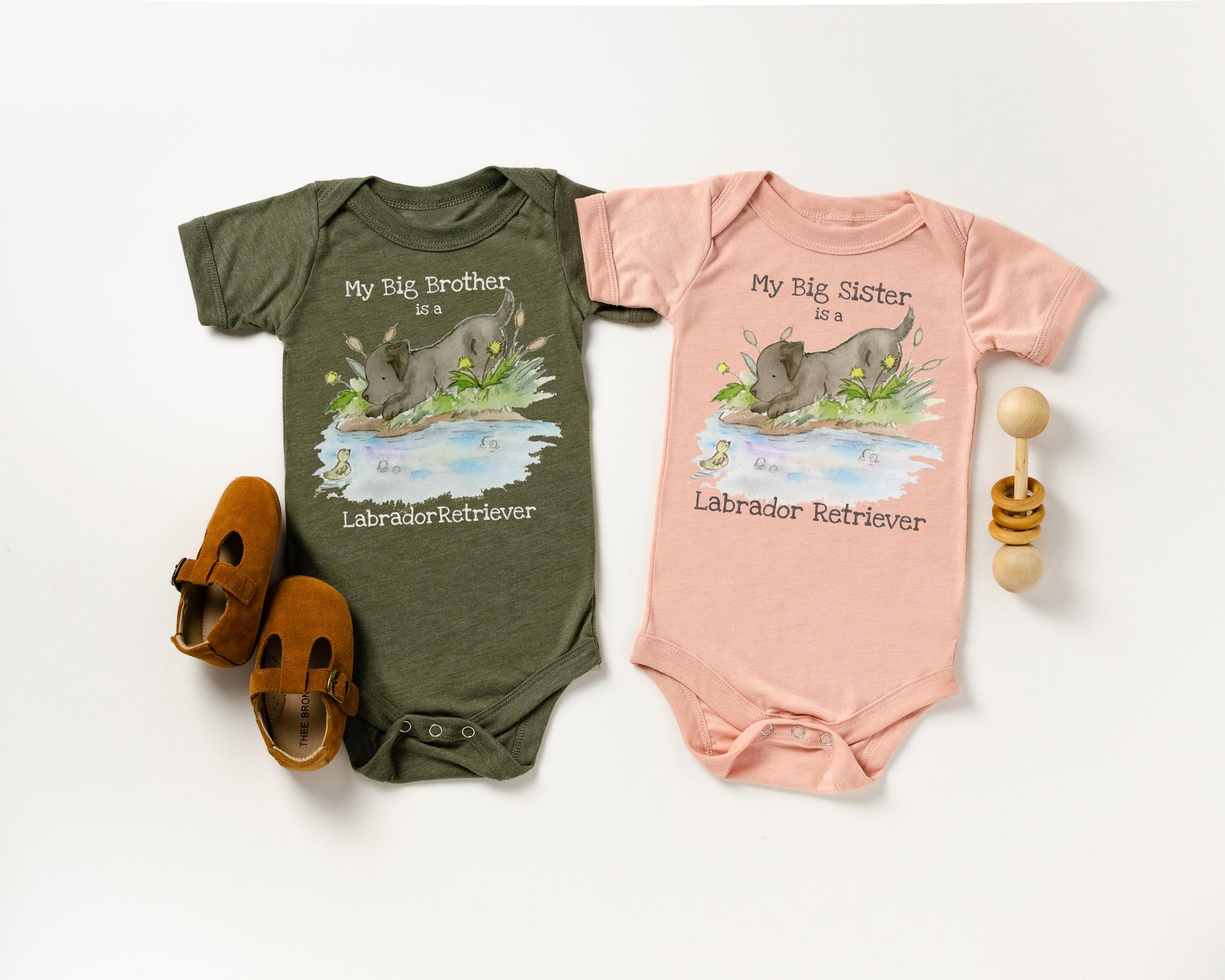 Two baby onesies, one green and one pink, with illustrations of black lab dogs and text on a white background.