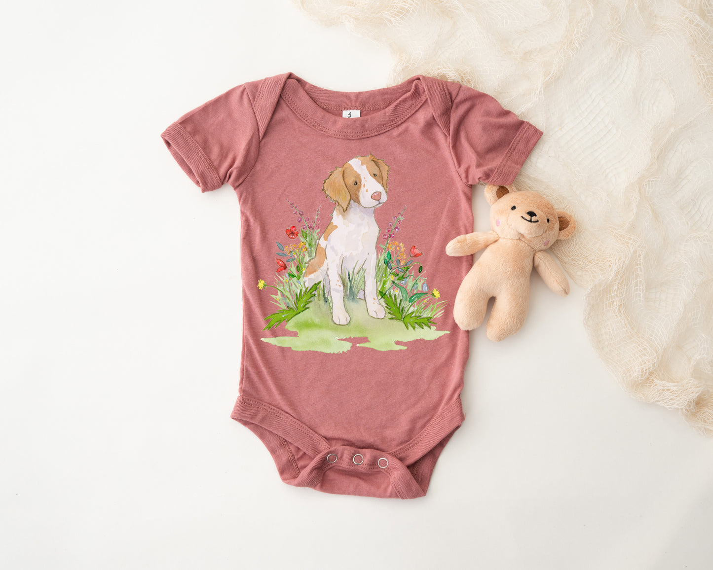 Pink baby onesie with a dog and floral design on a white surface with a teddy bear.