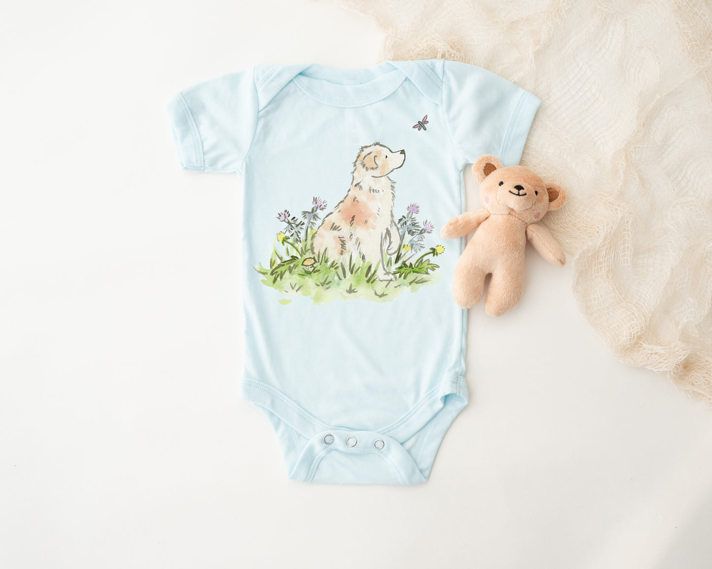 Light blue baby onesie with a dog and flower design, accompanied by a teddy bear on a white background.