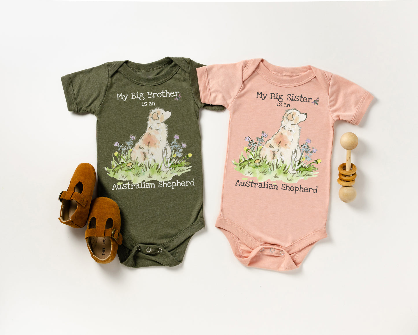 Two baby onesies, one green and one pink, with Australian Shepherd illustrations and text on a white background.