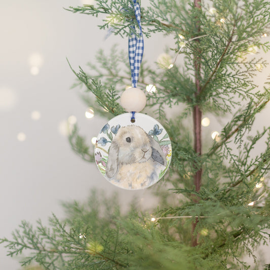 White ceramic ornament with blue ribbon and wooden bead with a painting of a tan and brown blue tort Holland Lop rabbit and colorful wildflowers.