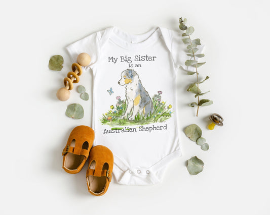 White baby bodysuit with a cute blue merle Australian shepherd and the words "my big sister is an Australian shepherd" on them.
