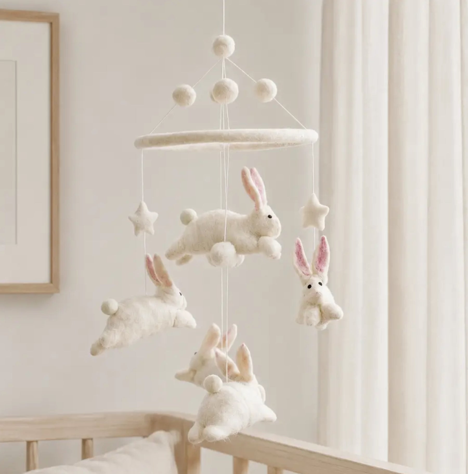 A soft white felt baby mobile with bunny and star shapes hanging in front of a crib in a bright nursery.