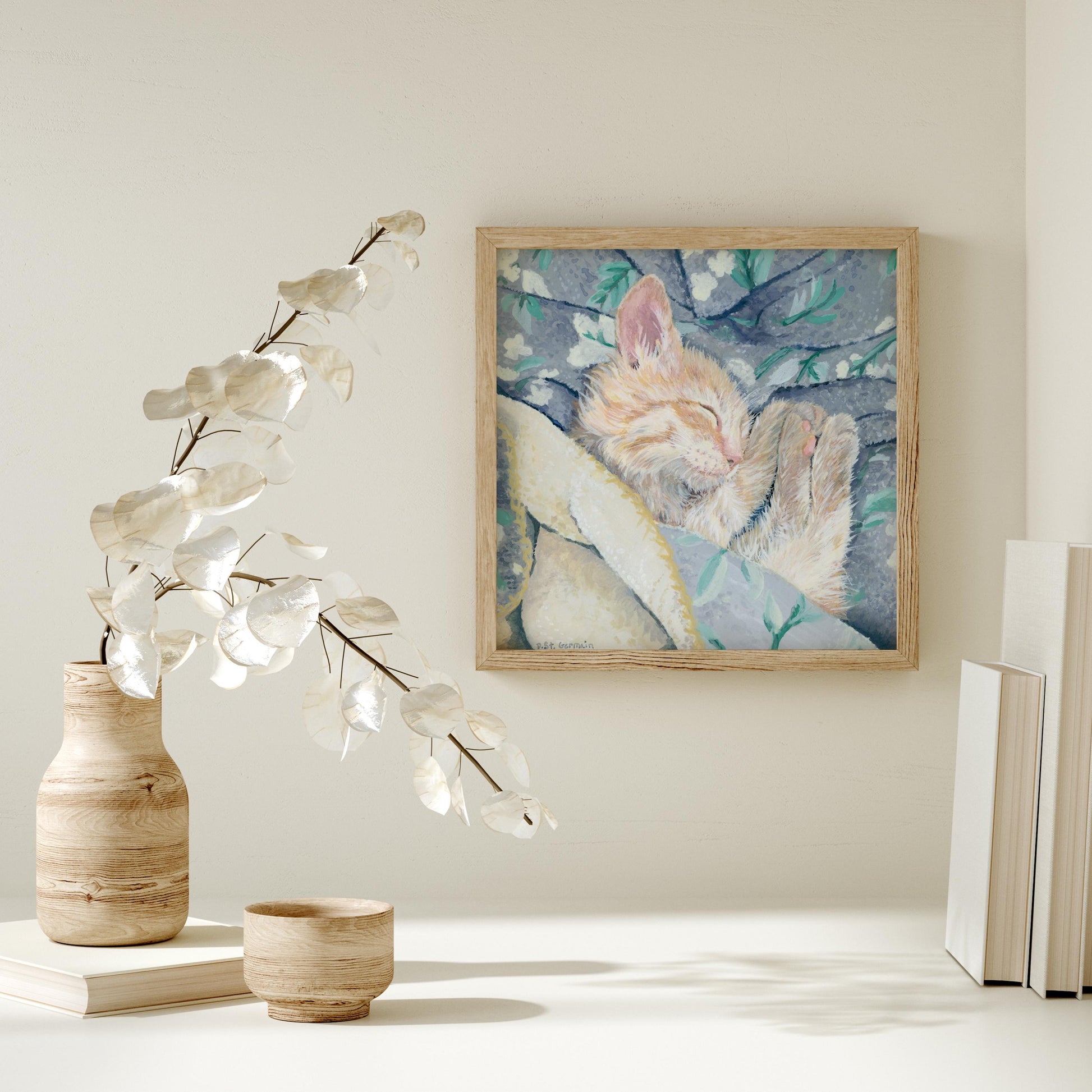 Framed gouache painting of an orange ginger kitten taking a nap in a gray and teal floral blanket.