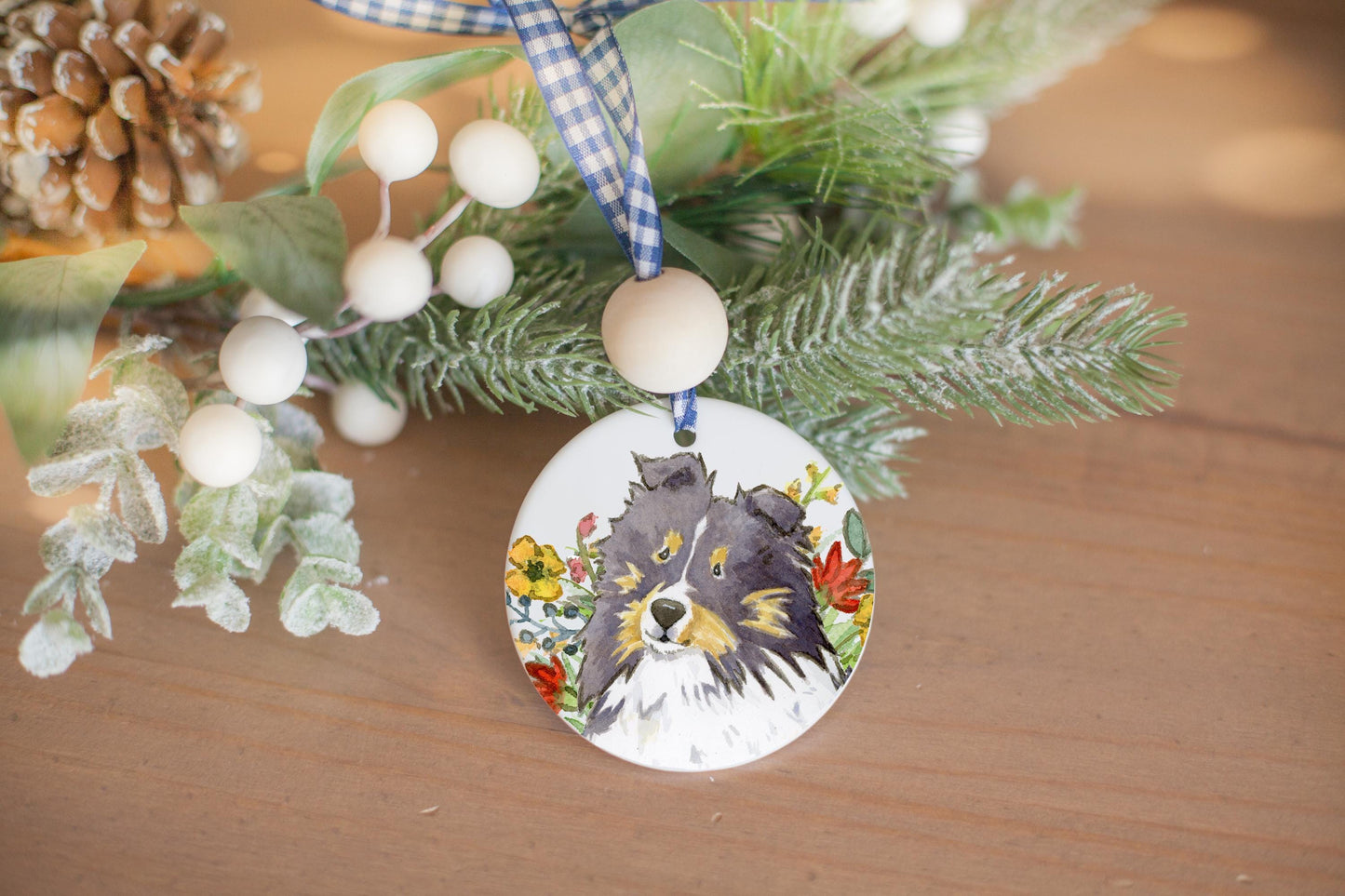 Tri color sheltie and colorful wildflowers on a ceramic ornament with holiday branches in te background.
