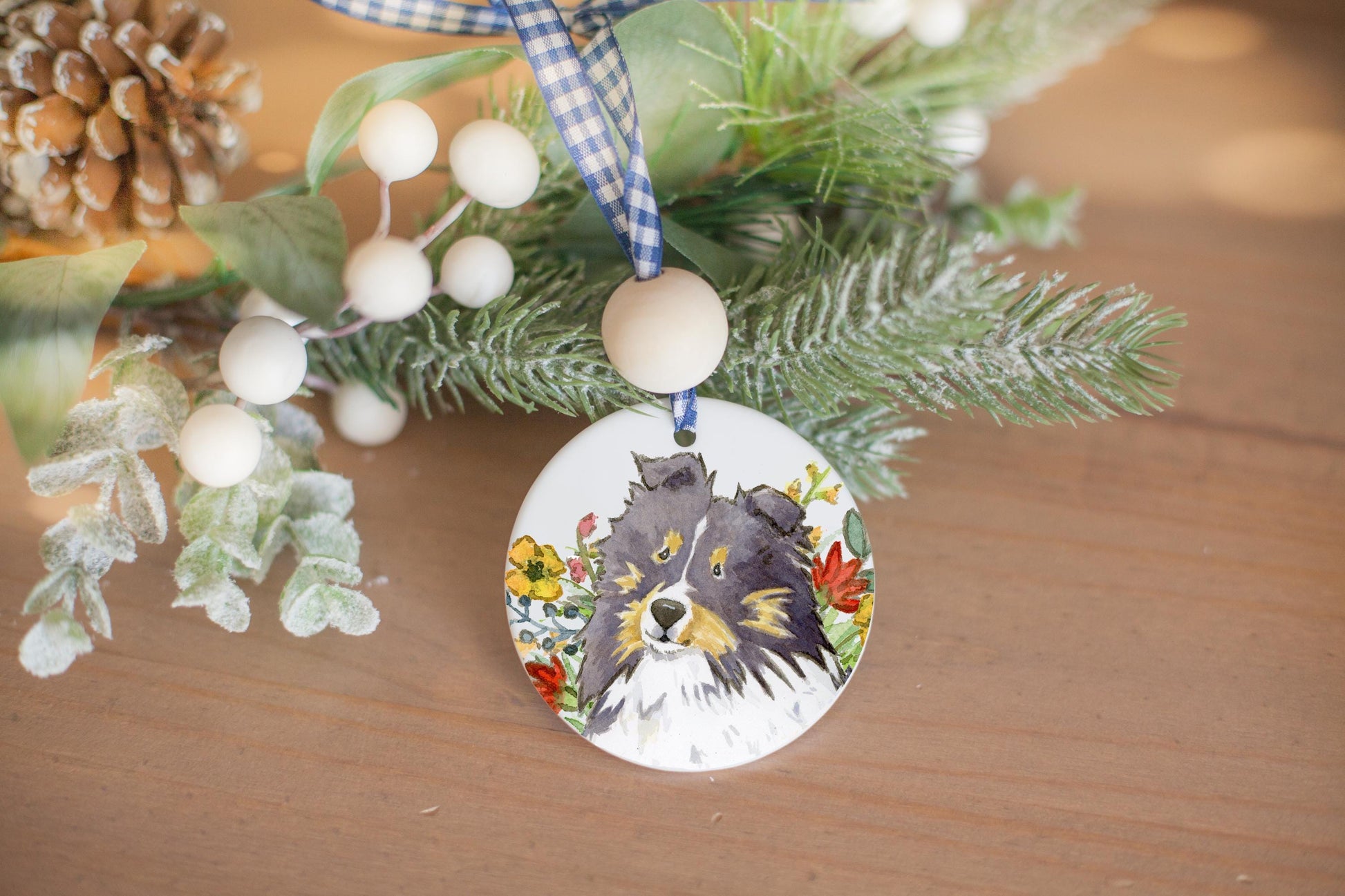 Tri color sheltie and colorful wildflowers on a ceramic ornament with holiday branches in te background.