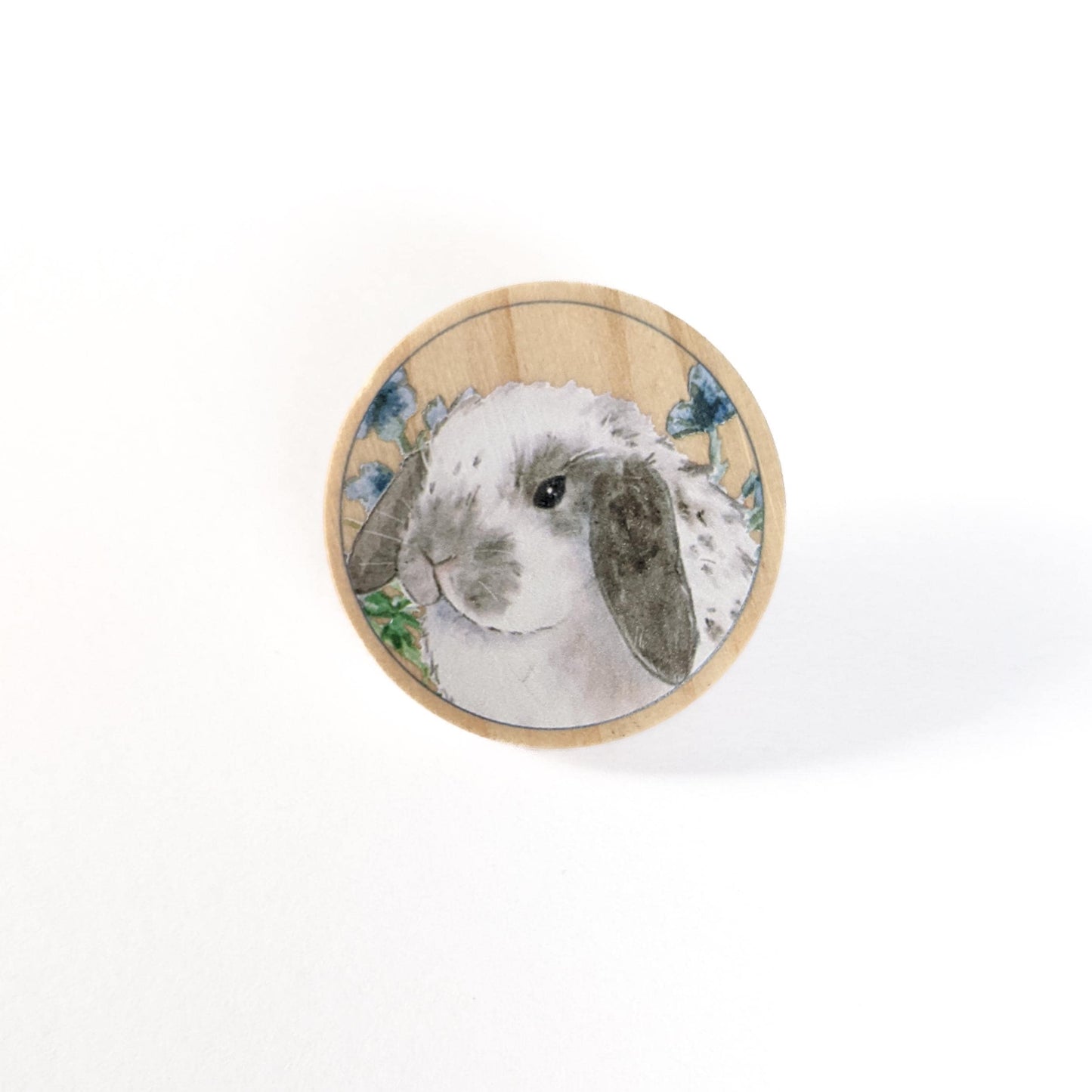 Round wooden pin with a gray and white spotted holland lop bunny and flowers on it.