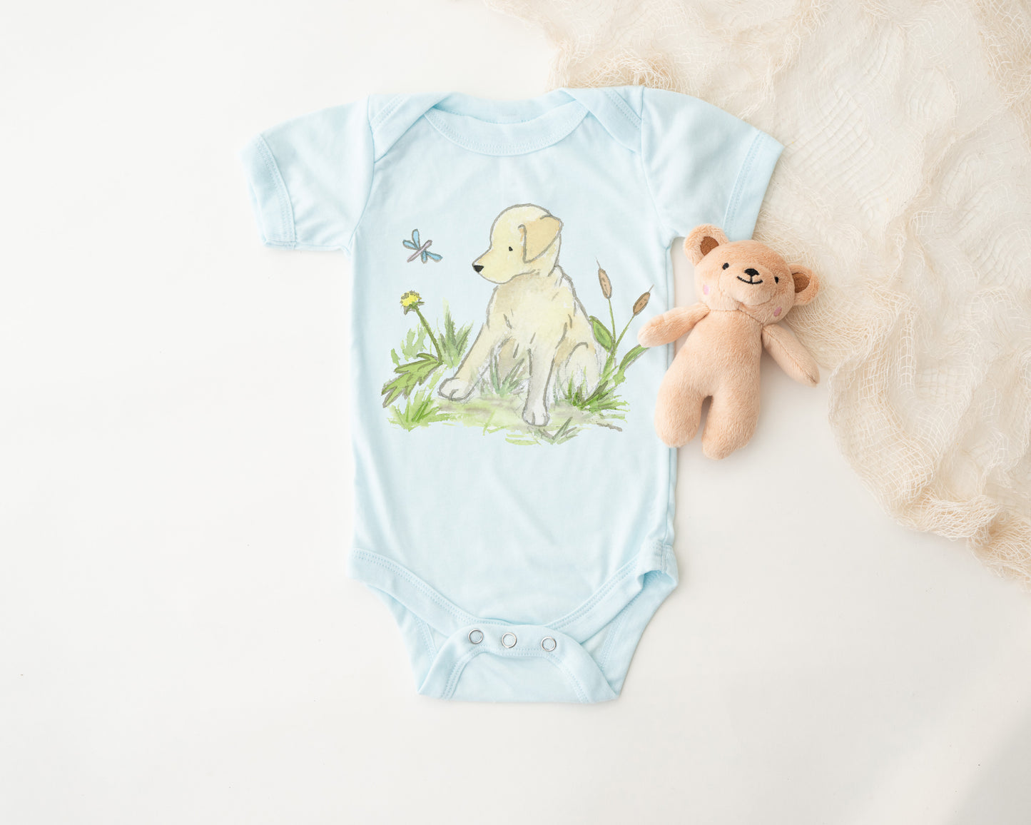 Light blue baby onesie with a dog illustration on a white surface with a teddy bear.