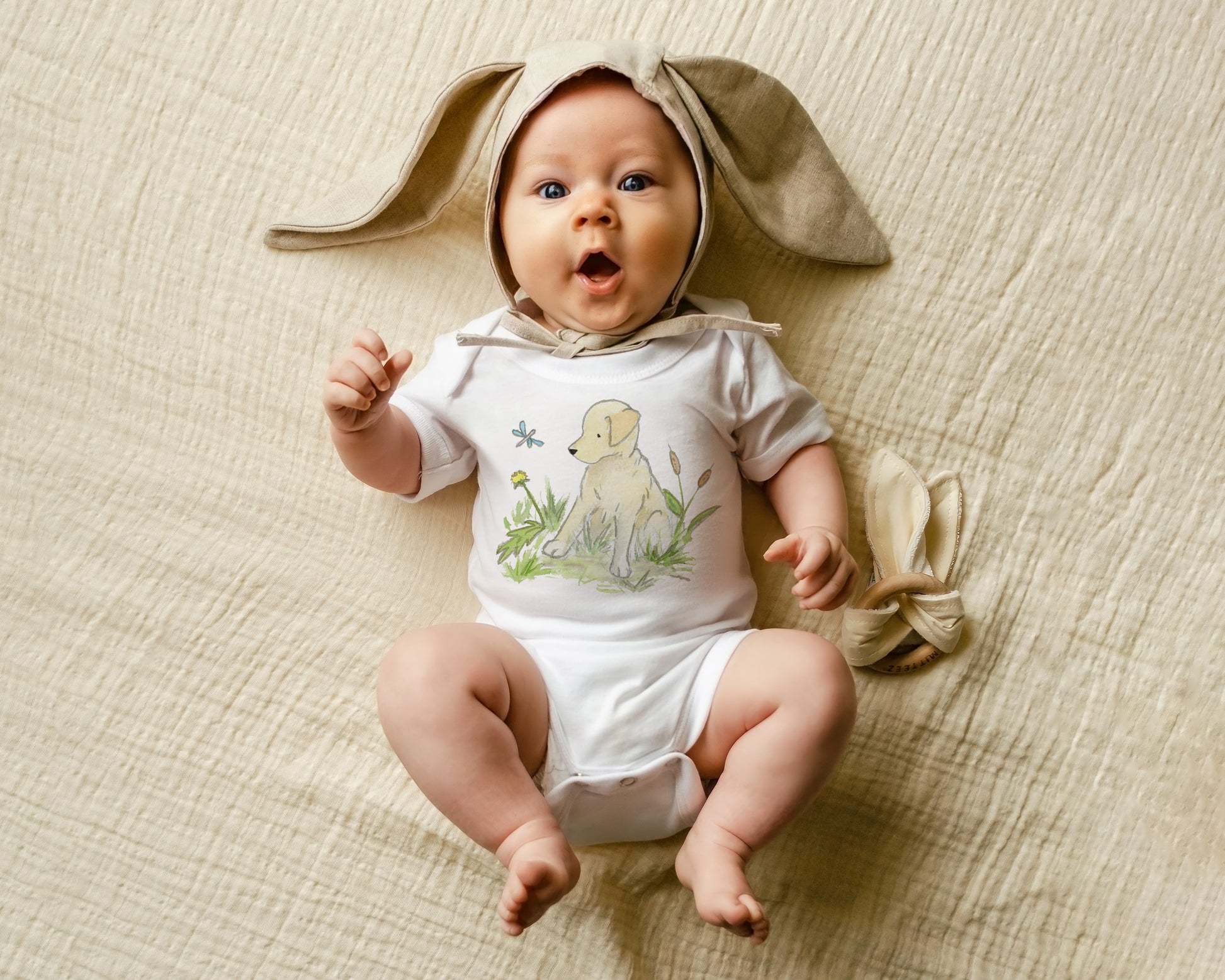 Baby wearing a bunny costume with a white shirt with a yellow lab on it and white pants on a beige textured surface